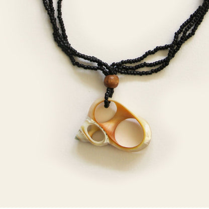 Closeup of handmade necklace with black seed beads, wooden accents, and a natural oval shell pendant with mini shell charms — by Nayabijou.