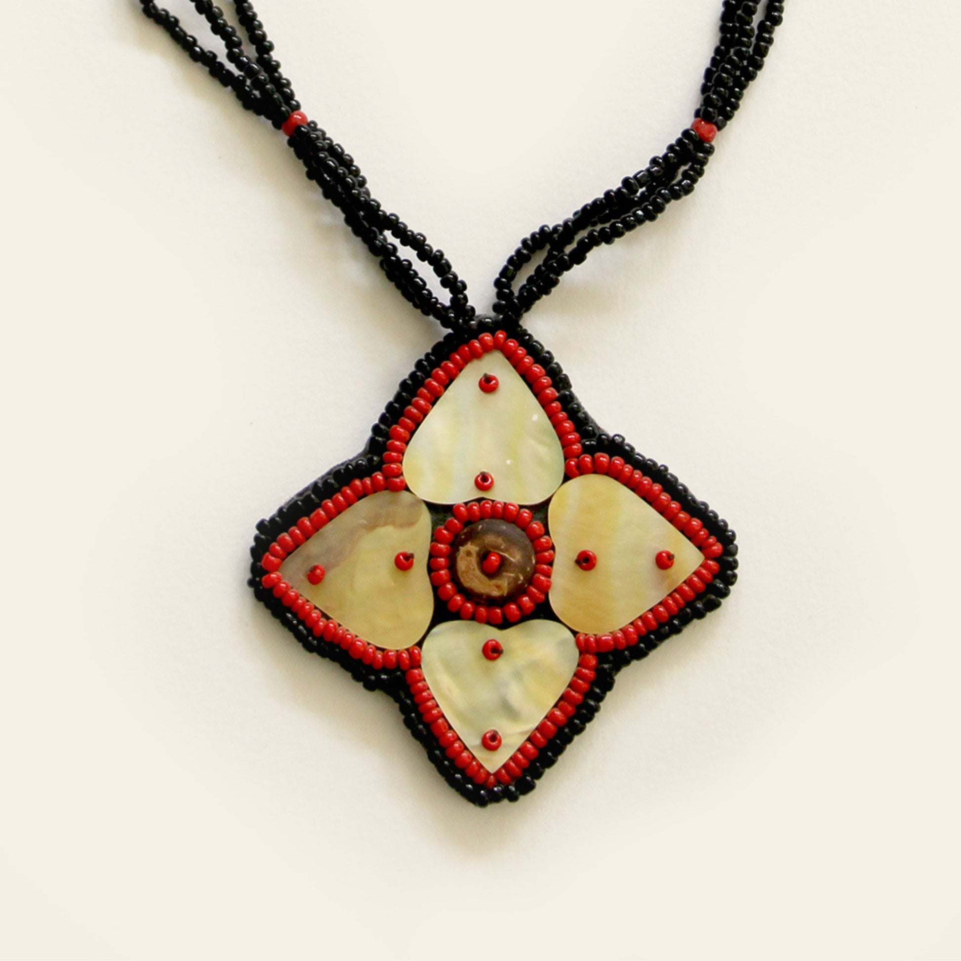 Closeup of handmade necklace featuring a mother-of-pearl flower pendant with red bead detailing, strung on triple black seed bead strands — by Nayabijou.