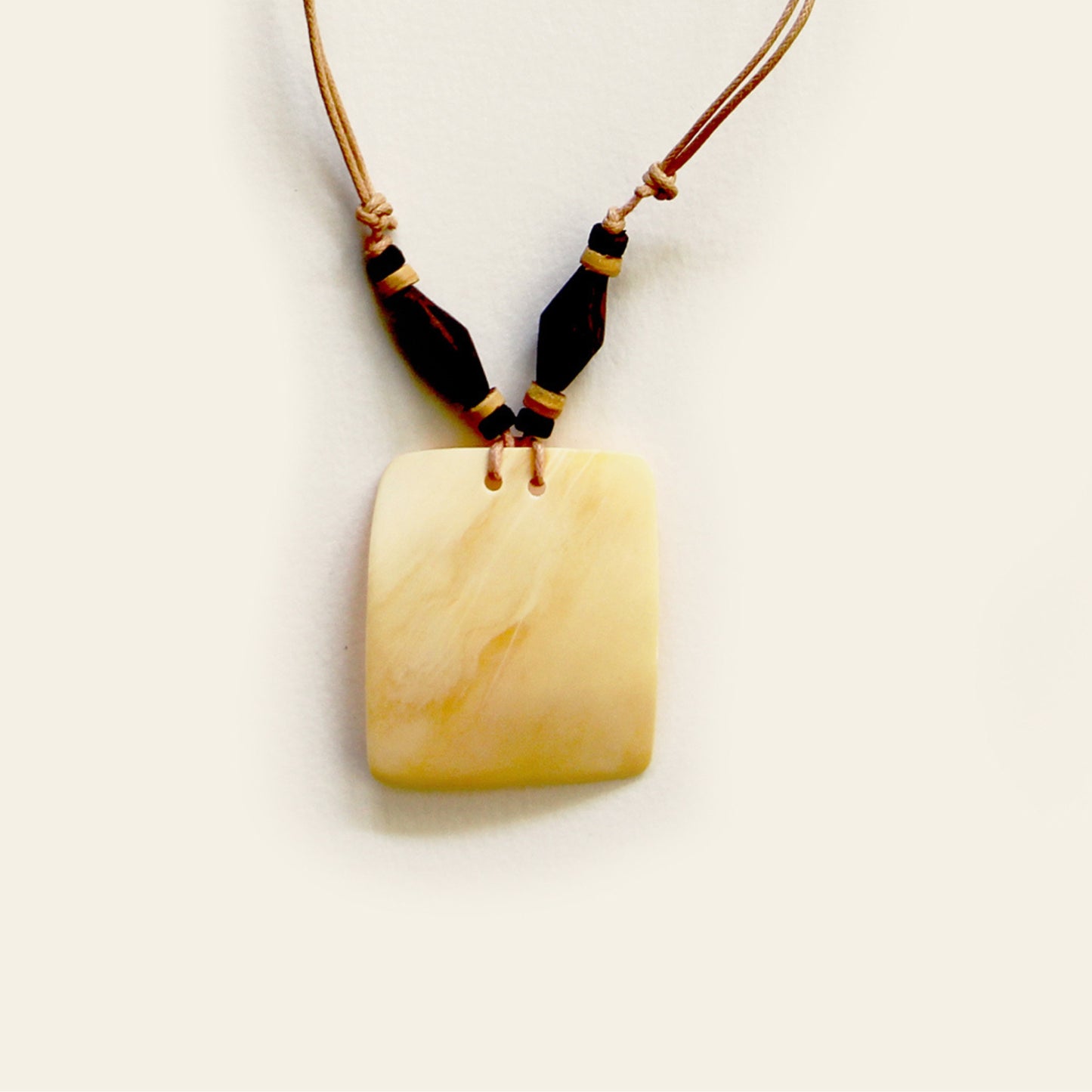 Closeup of handmade necklace with a tan leather cord, dark wood beads, and a square-shaped beige shell pendant — by Nayabijou.