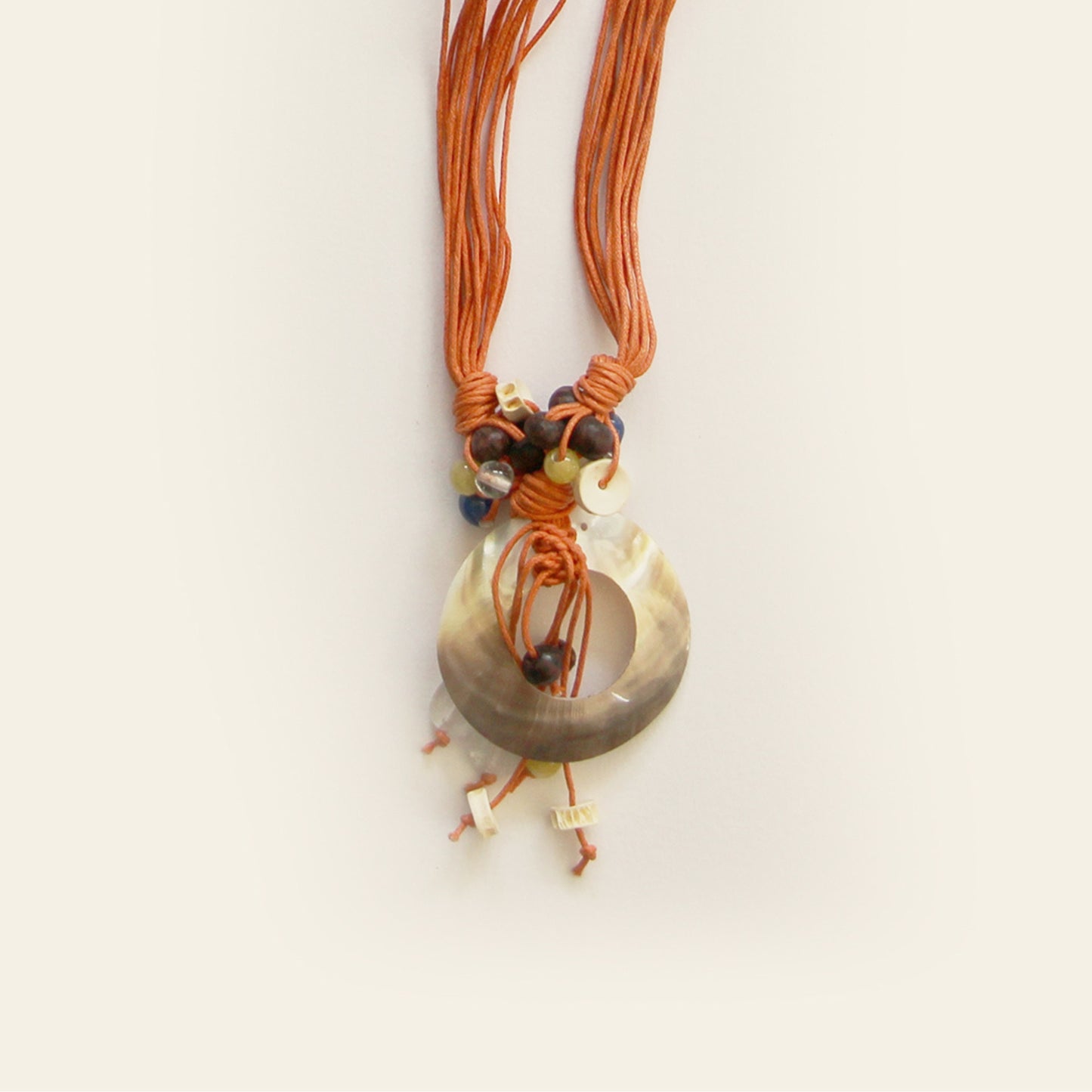 Closeup of a handmade necklace with burnt orange cords, a shell pendant, and small natural beads – by Nayabijou.