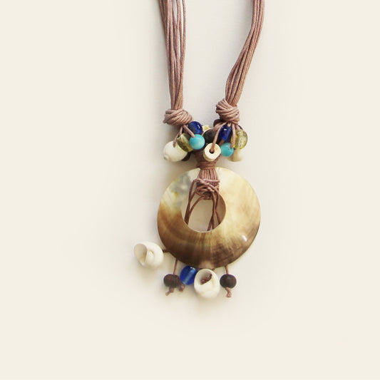 Closeup of a handmade necklace with taupe cords, a round shell pendant, and a cluster of natural beads – by Nayabijou.