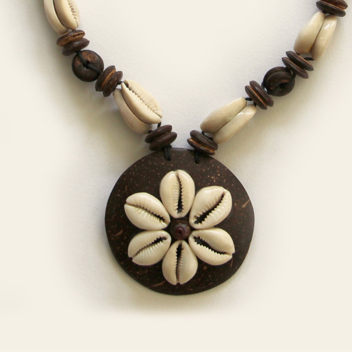 Closeup of handmade necklace with natural cowrie shells arranged on a dark round pendant, paired with wood and shell beads on a black cord — by Nayabijou.