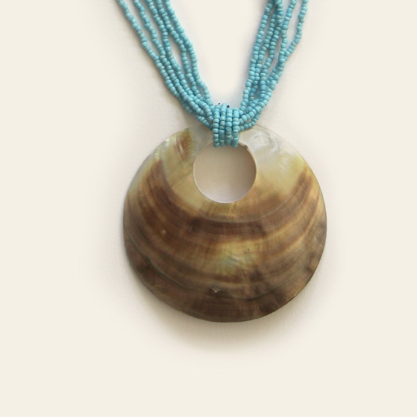 Closeup of handmade necklace with a large round shell pendant and layered turquoise seed bead strands — by Nayabijou.