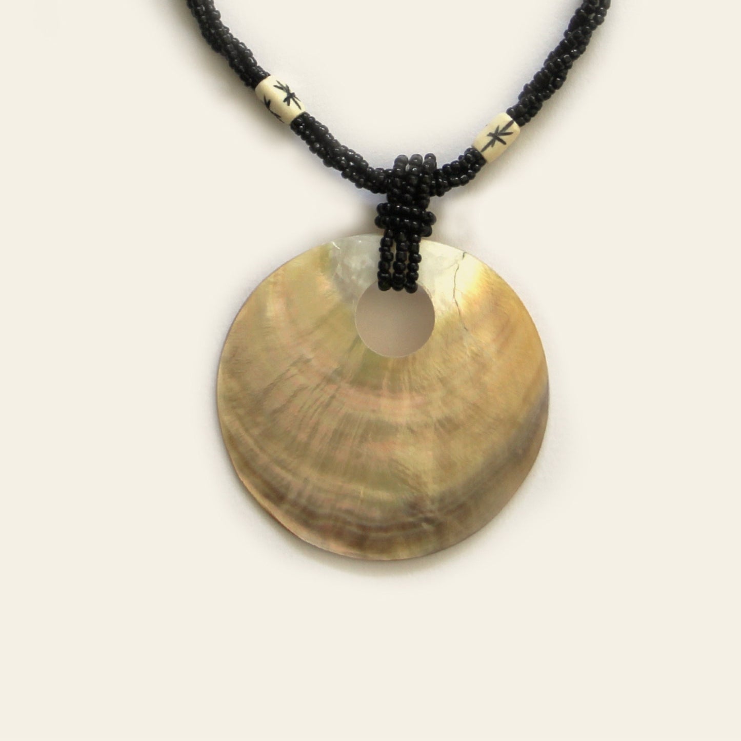 Closeup of handmade necklace with black seed beads, ivory accents, and a large golden-brown round shell pendant — by Nayabijou.