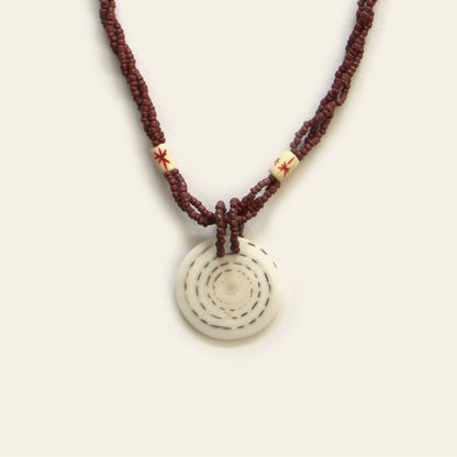 Handmade necklace with brown seed beads, dark wooden accents, and a round spiral-patterned shell pendant — by Nayabijou.