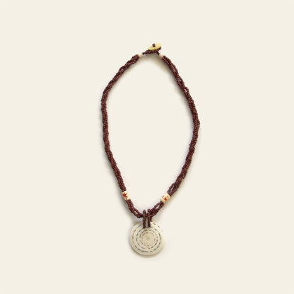Handmade necklace with brown seed beads, dark wooden accents, and a round spiral-patterned shell pendant — by Nayabijou.