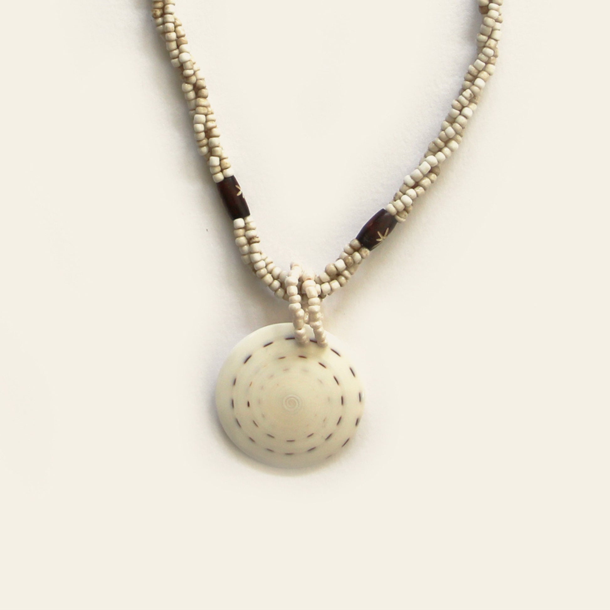 Closeup of handmade necklace with sand seed beads, dark wooden accents, and a round spiral-patterned shell pendant — by Nayabijou.