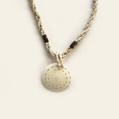 Closeup of handmade necklace with sand seed beads, dark wooden accents, and a round spiral-patterned shell pendant — by Nayabijou.