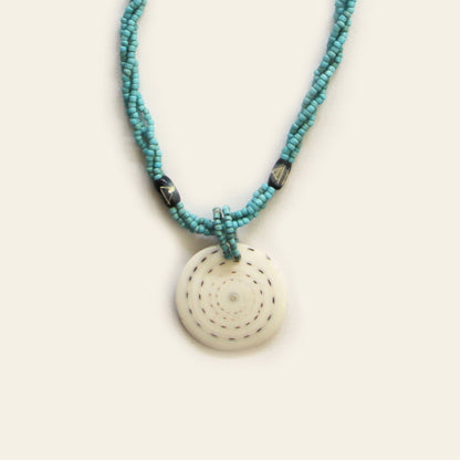 Closeup of handmade necklace with turquoise seed beads, dark wooden accents, and a round spiral-patterned shell pendant — by Nayabijou.