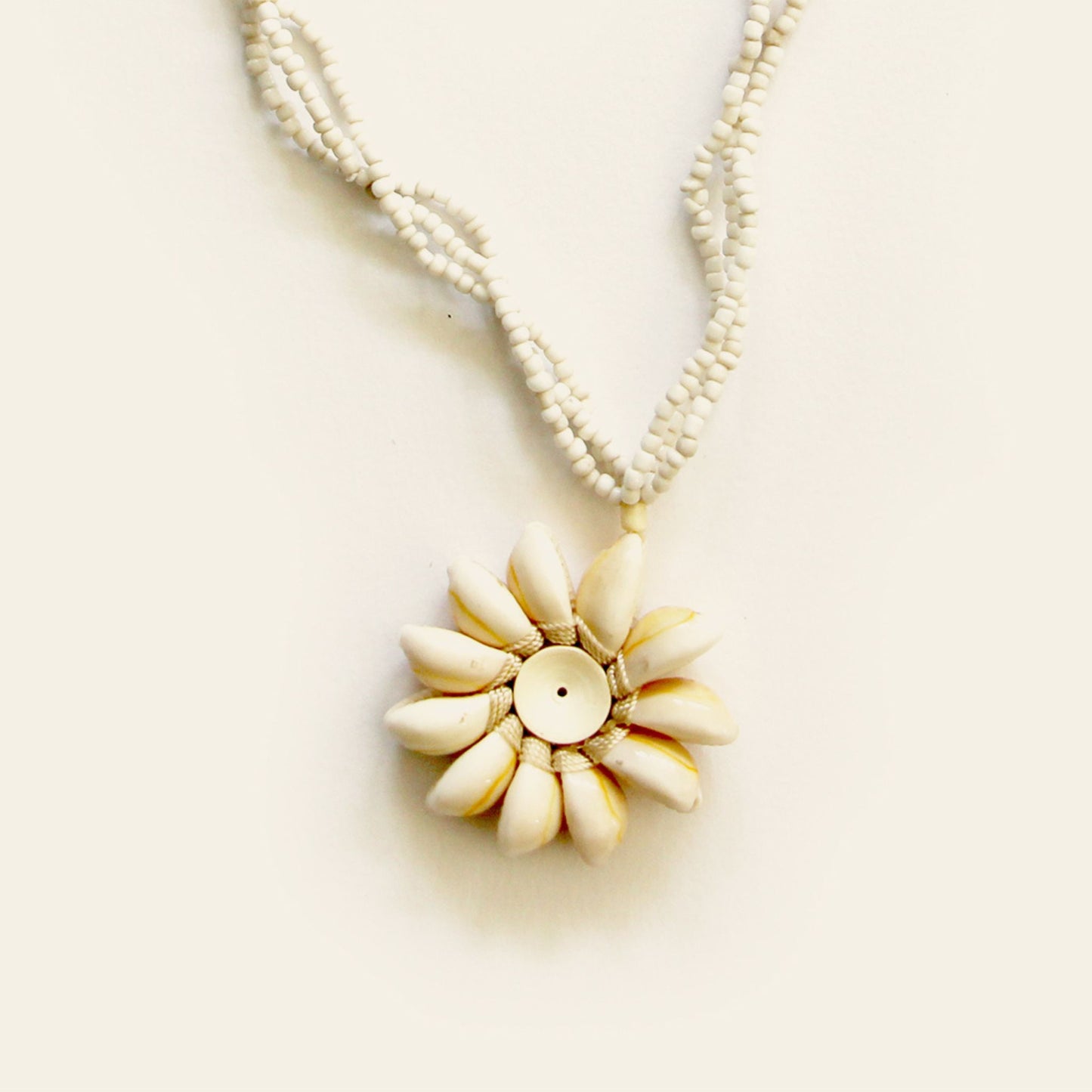 Closeup of handmade necklace with white seed bead strands and a central flower pendant made of natural shells arranged in a sunburst pattern — by Nayabijou.