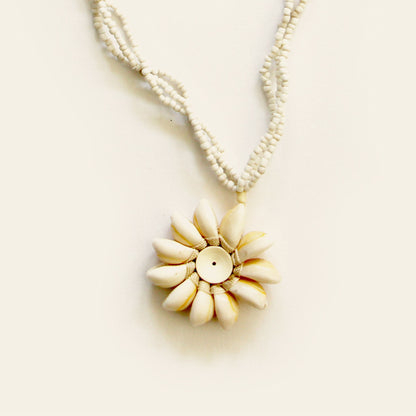 Closeup of handmade necklace with white seed bead strands and a central flower pendant made of natural shells arranged in a sunburst pattern — by Nayabijou.
