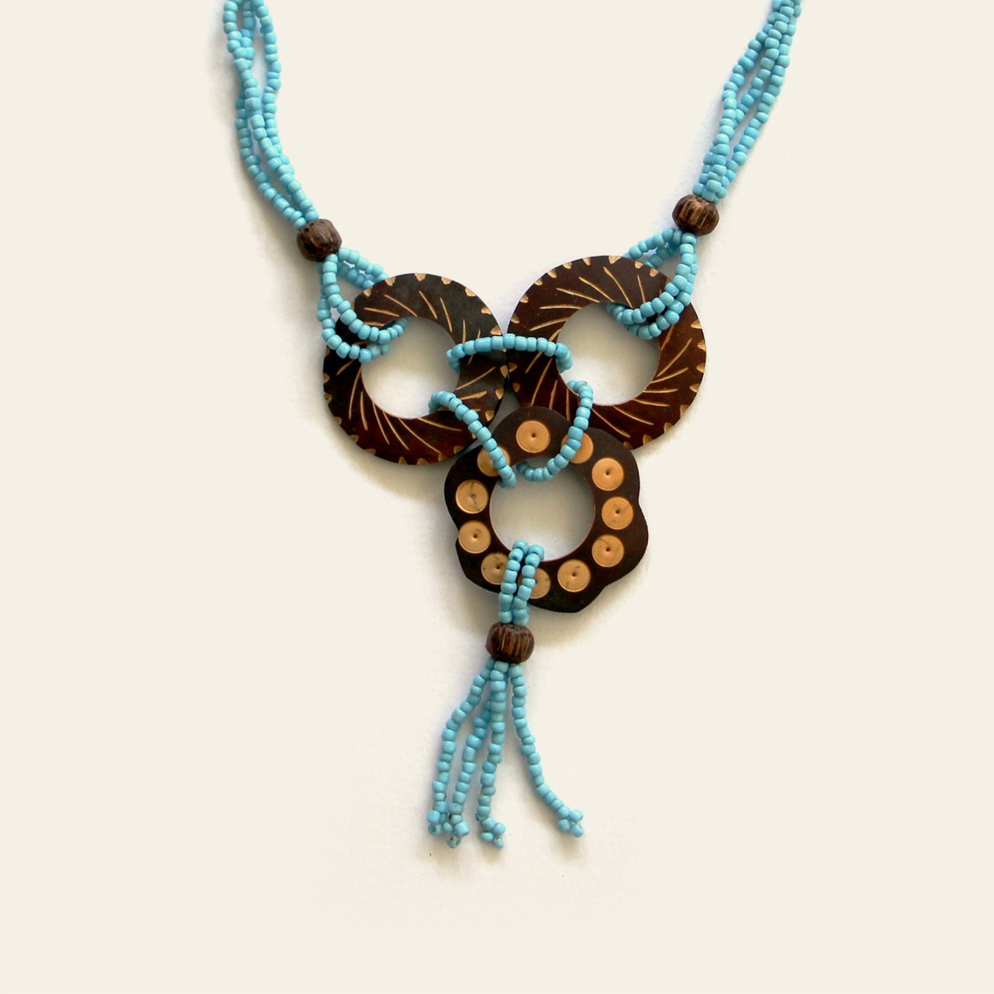 Closeup of a handmade necklace featuring Turquoise seed beads and carved coconut shell loops linked together with beaded knots, finished with a tasseled drop — by Nayabijou.
