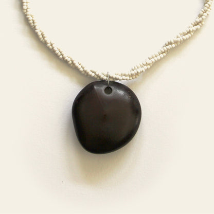 Closeup of handmade necklace with off-white seed beads and a smooth black wooden pendant, designed by Nayabijou.