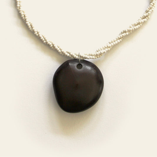 Closeup of handmade necklace with off-white seed beads and a smooth black wooden pendant, designed by Nayabijou.